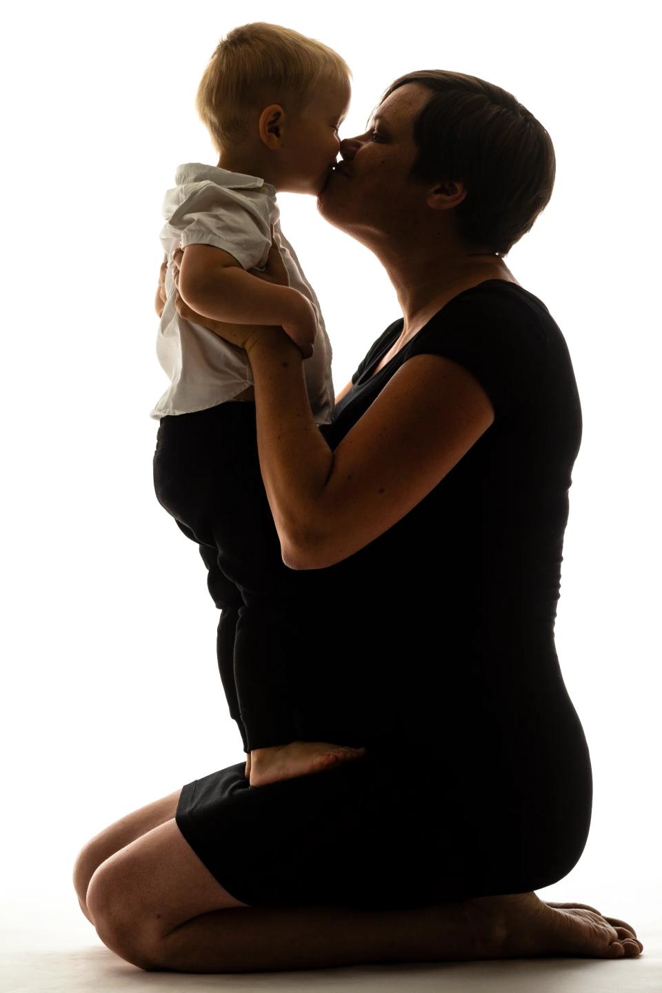 Gravid fotografering i sort/hvid med silhuet af en knælende, gravid mor, der kysser sit lille barn, holdt tæt ind til sig.