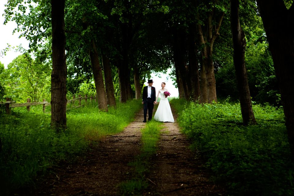 Bryllupsfoto: Et brudepar går hånd i hånd på en grussti flankeret af grønne træer og frodig vegetation i smukt, blødt lys.
