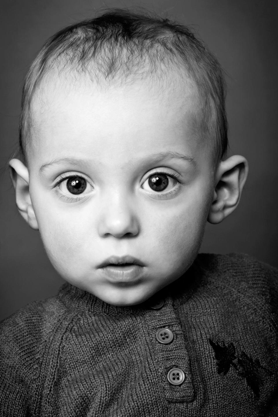 Sort/hvid portrætfoto af nysgerrig baby der ser direkte i kameraet med store øjne i blødt lys. Børn