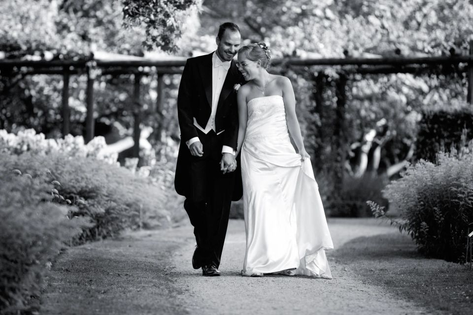 Bryllupsfoto i sorthvid af smilende brudepar, der holder i hånd og går på havesti i blødt sidelys.