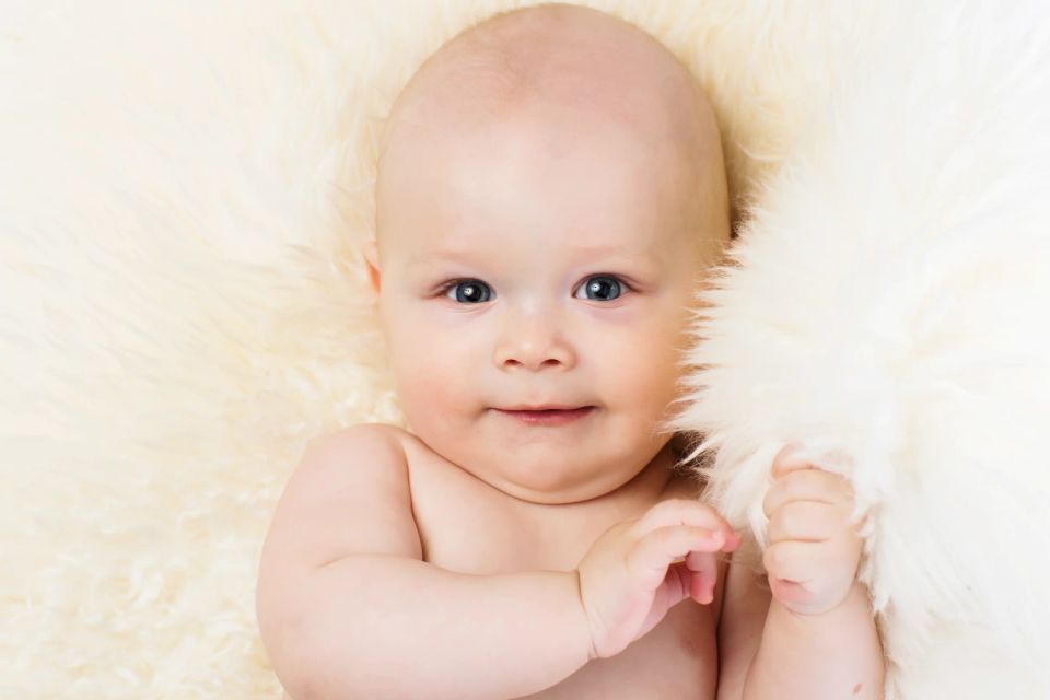 Glad baby ligger på hvidt lammeskind. Blød babyfotografering med varme, dæmpede farver og skarpt fokus.