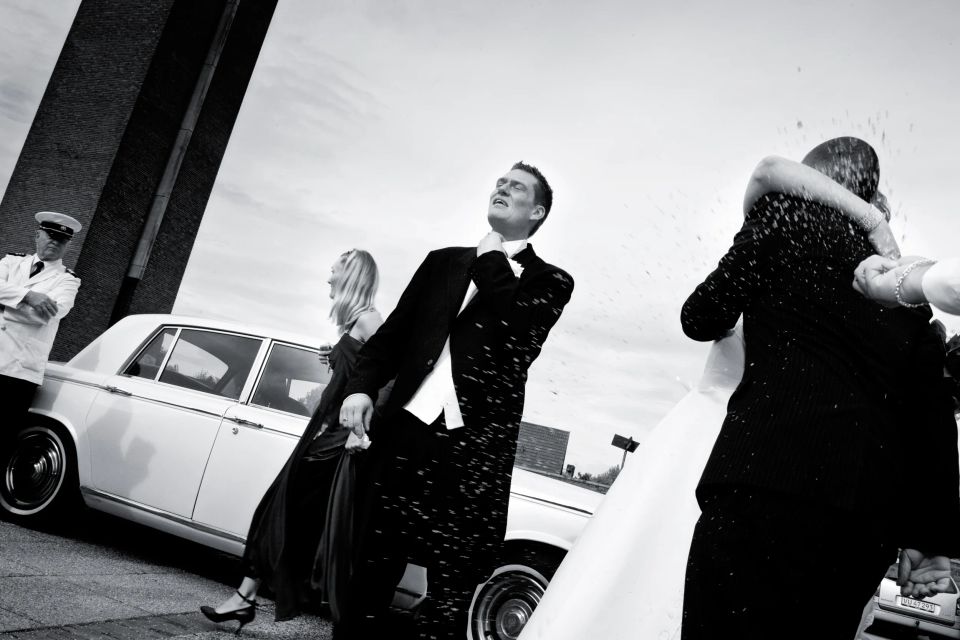 Bryllupsfoto i sorthvid af gæster, der kaster ris på brudeparret foran en hvid bil ved ankomst. Festlig stemning.