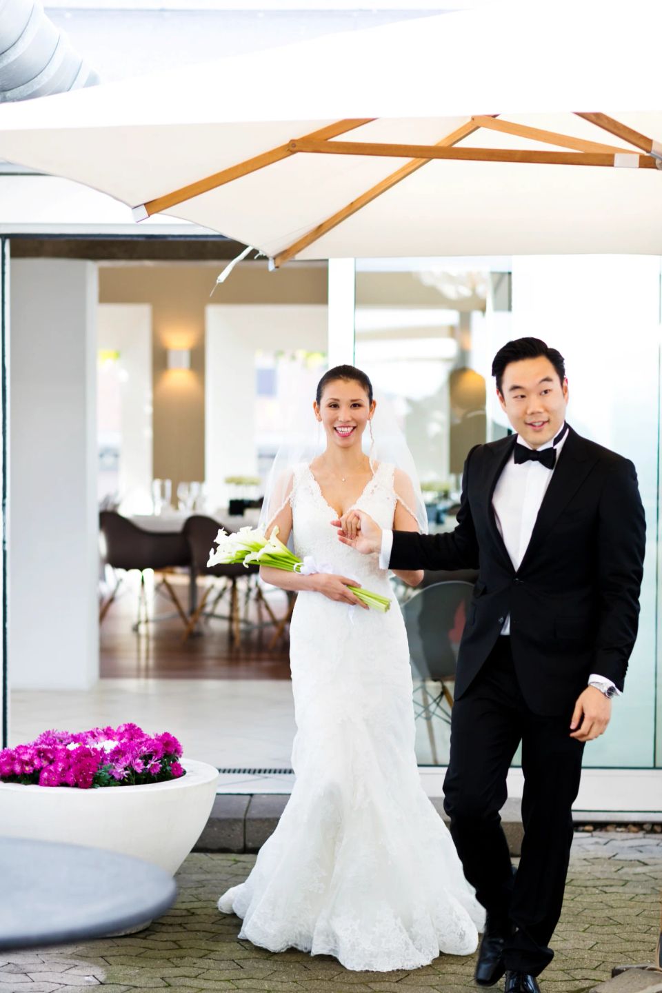 Bryllupsfoto: Smilende nygift par ankommer hånd i hånd til receptionen, bruden holder en buket hvide kalaer.