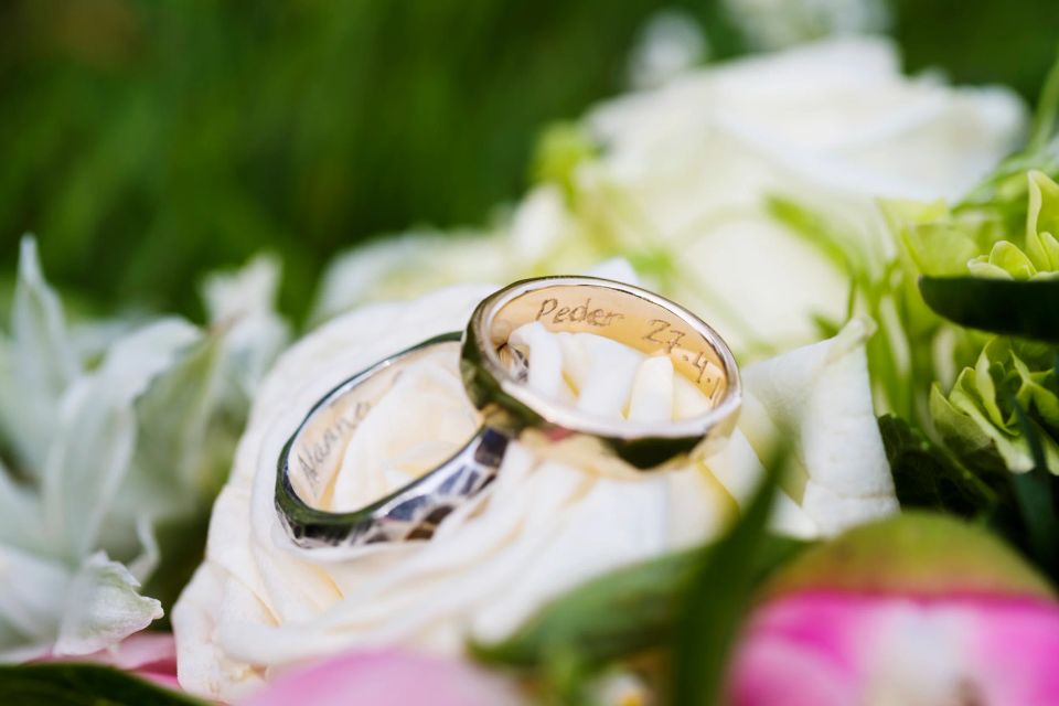 To vielsesringe med indgraverede navne og dato, placeret i en smuk bryllupsbuket med hvide og grønne nuancer.
