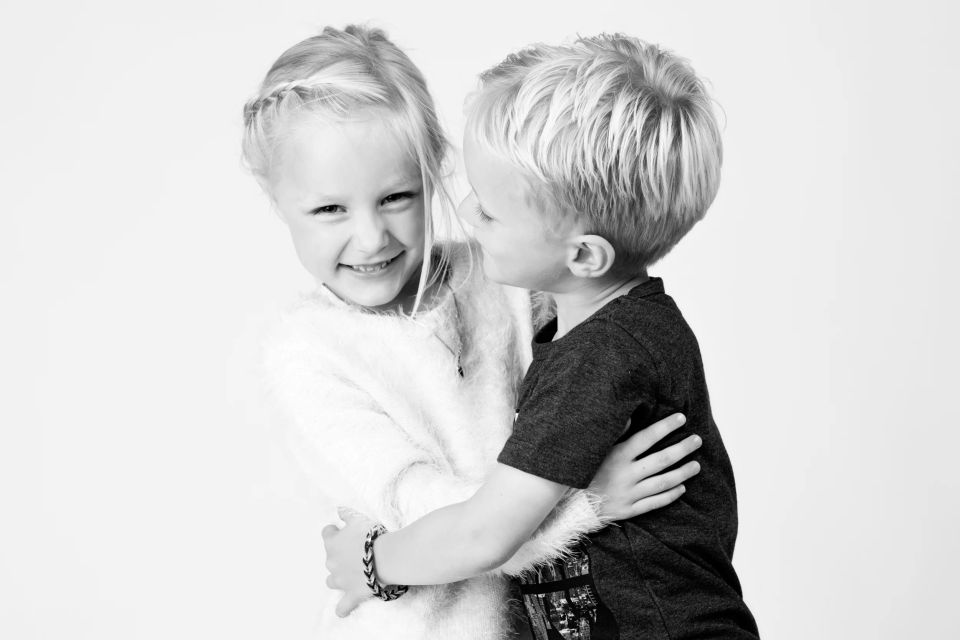 Børneportræt i sort/hvid af smilende pige som krammer sin lillebror på en lys baggrund. Søskendekærlighed