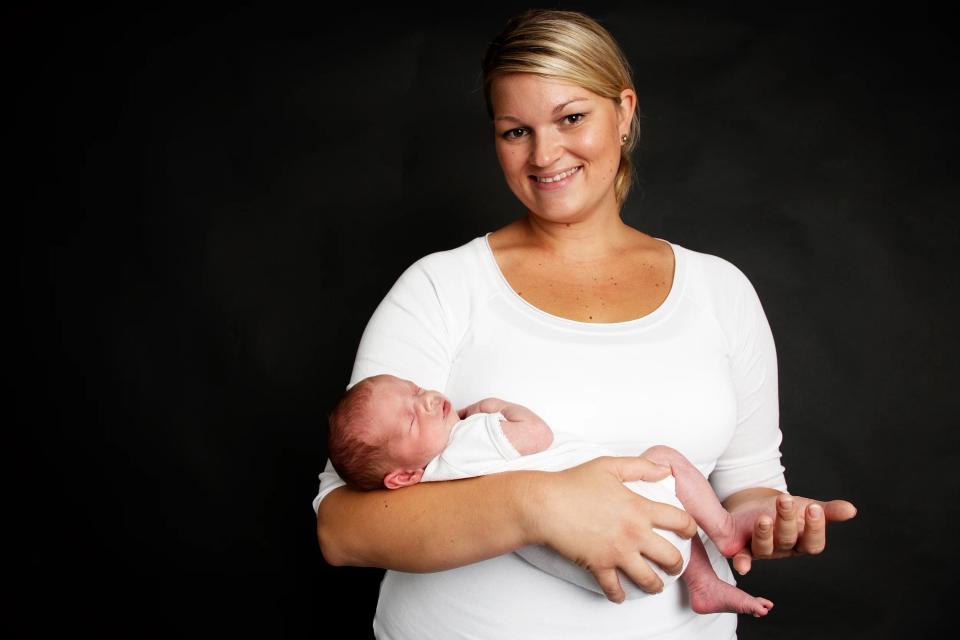 Glad mor smiler til kameraet, mens hun holder sin sovende nyfødte baby. En rørende babyfotografering.