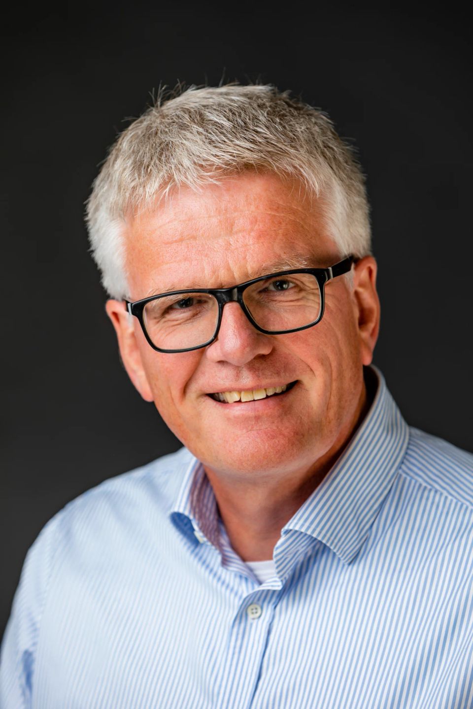 Nærbillede, portrætfoto af smilende mand med gråt hår og sorte briller i naturligt lys mod mørk baggrund. Glade mundvinkel.