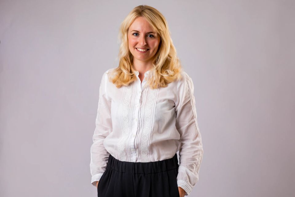 Portrætfoto af smilende kvinde med langt, blondt hår i hvid skjorte og sorte bukser mod grå baggrund.