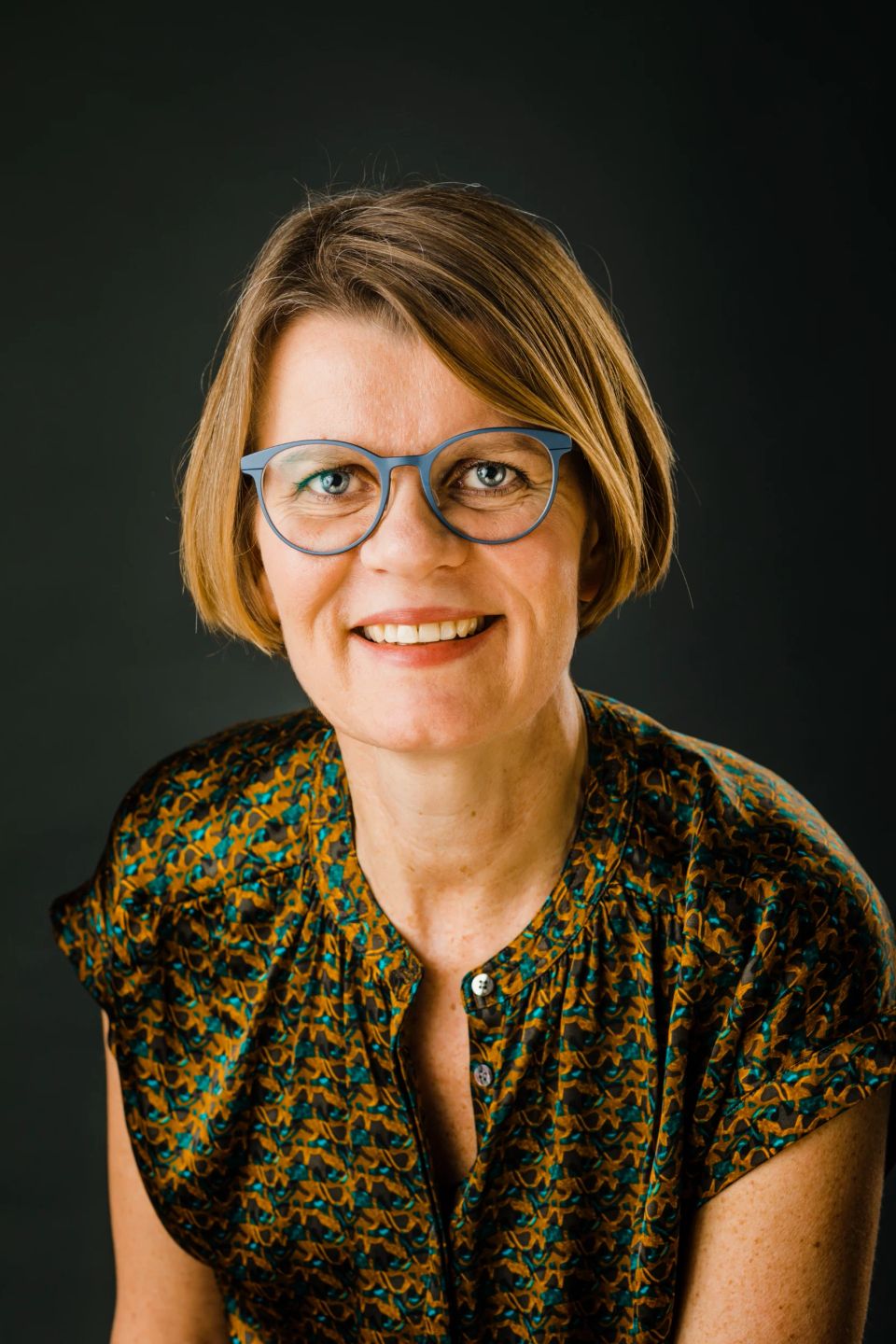 Portrætfoto af smilende kvinde med kort, lyst hår og blå briller, lys sat mod mørk baggrund.