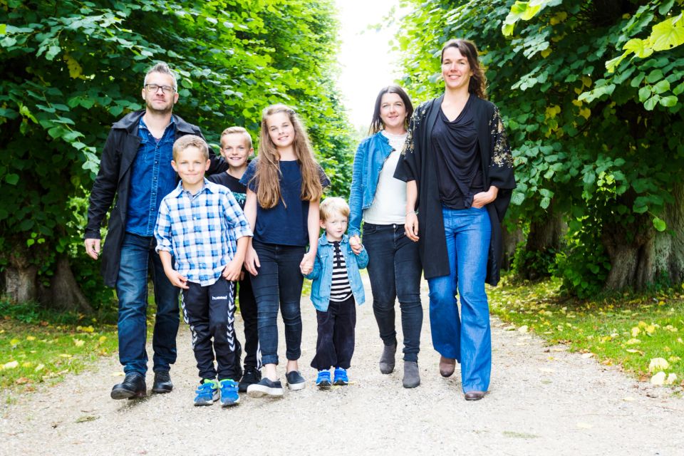 Familiefoto af syv glade personer i forskellige aldre, der går hånd i hånd ad en sti omkranset af grønne træer.