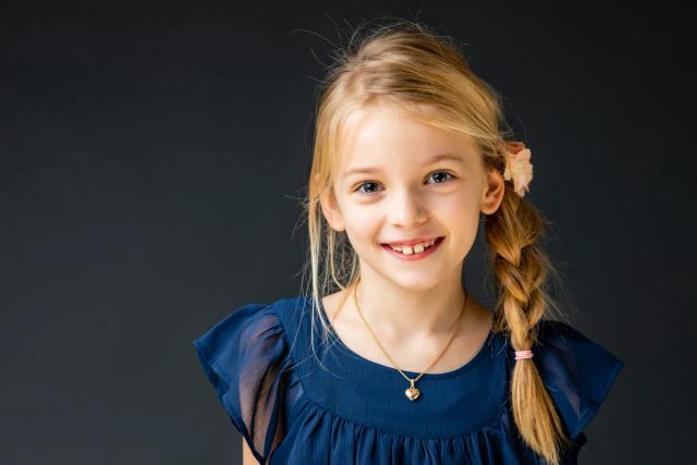Nærbillede af glad blond pige med smil og fletning, mørk baggrund. Et charmerende børnefoto i studiet.