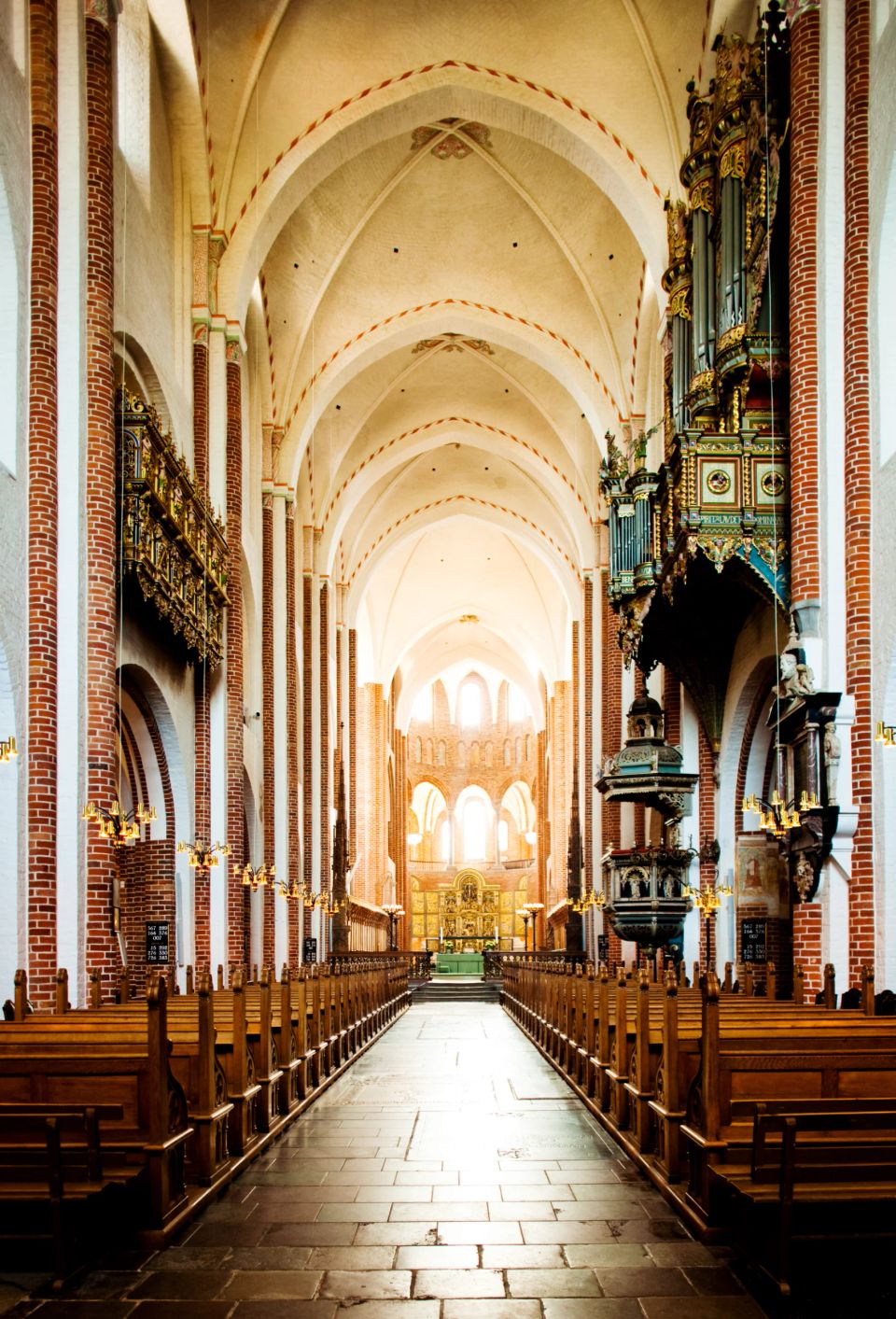 Historisk kirkerum fotograferet ned gennem midtergangen med rækker af træbænke, et stort gyldent orgel og lyst alter.