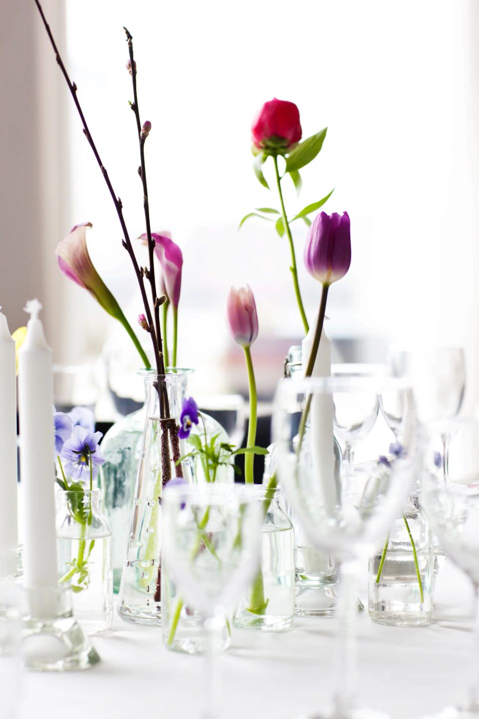 Festlig bryllupsborddekoration: Smukke blomster som lilla tulipaner og lyserøde kallaer i klare vaser på hvid dug.