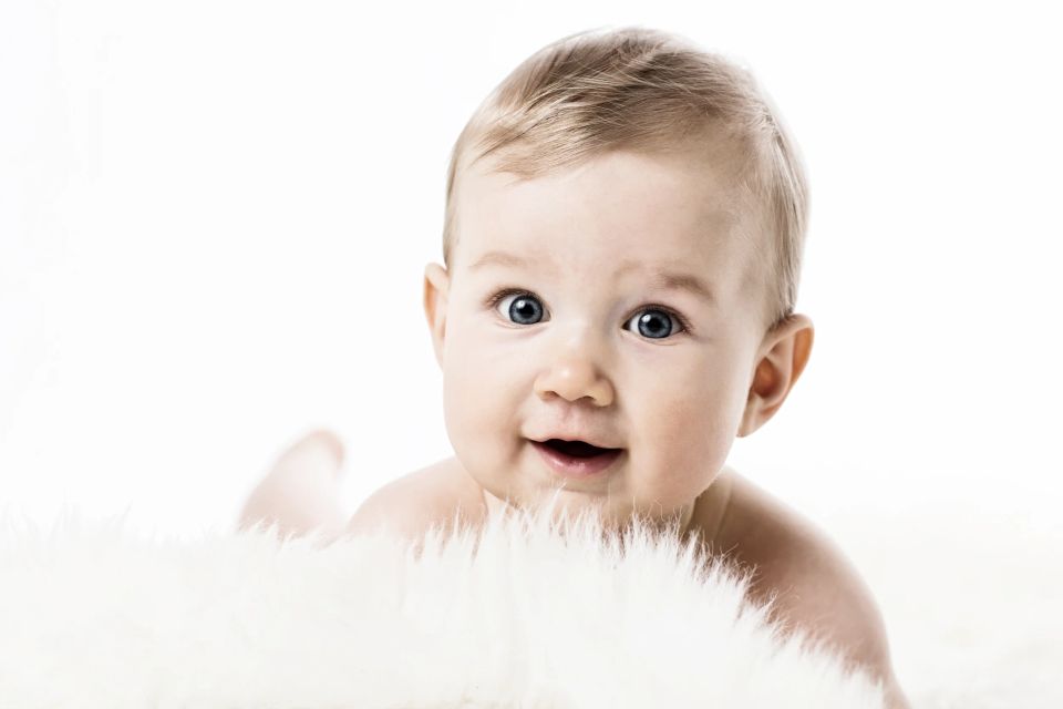 Glad baby med blå øjne og lyst hår ligger på maven, smilende på et hvidt lammefæld i blødt studielys. Babyfotografering.