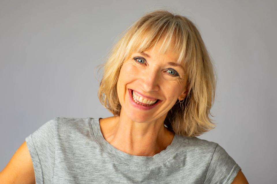 Portrætfoto af smilende blond kvinde med glade blå øjne i naturligt varmt lys mod en grå baggrund.