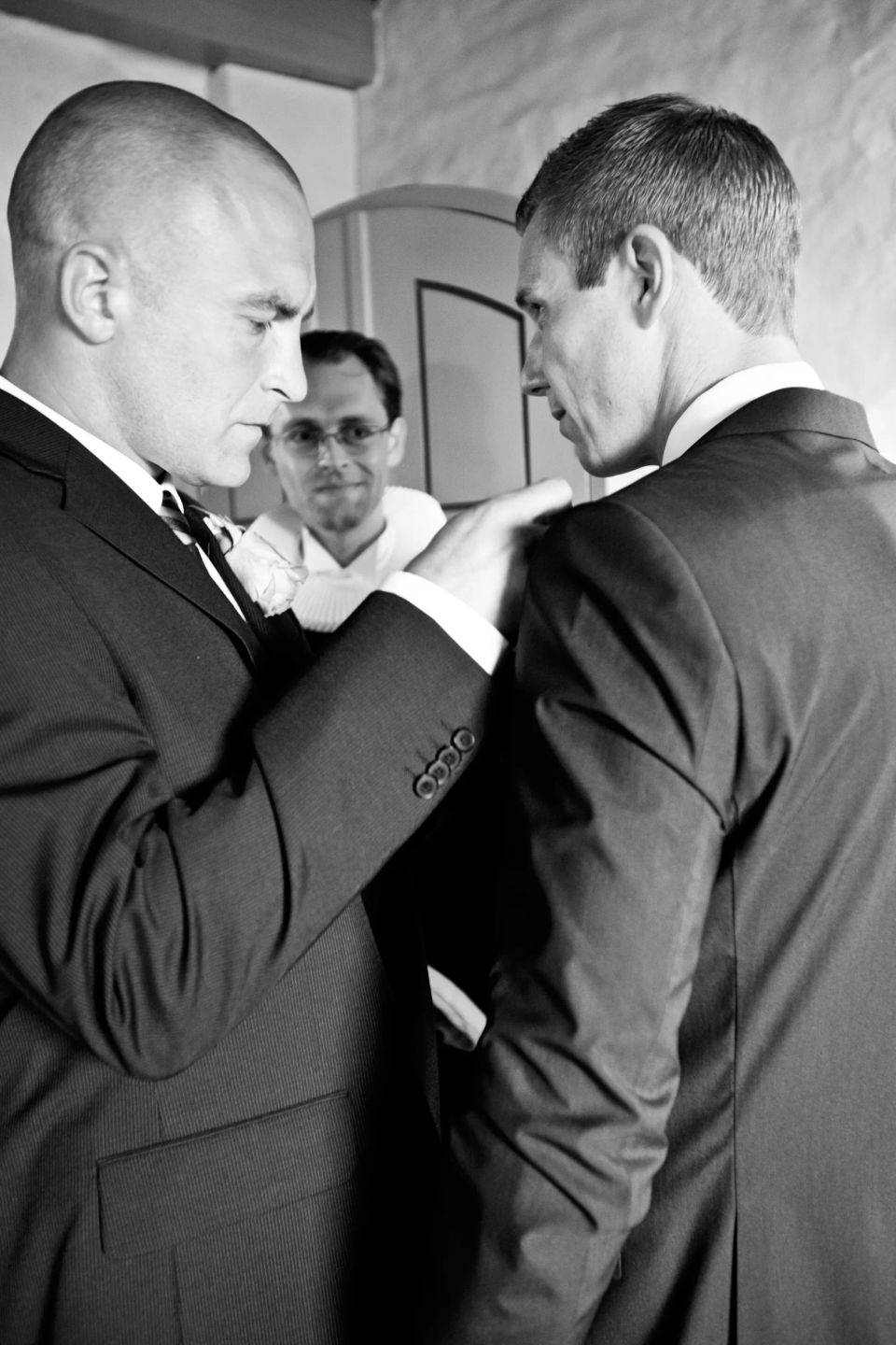 Bryllupsfoto af to gæster i sorte jakkesæt, den ene hjælper den anden med tøjet før bryllupsfesten. En tredje mand ses i b...