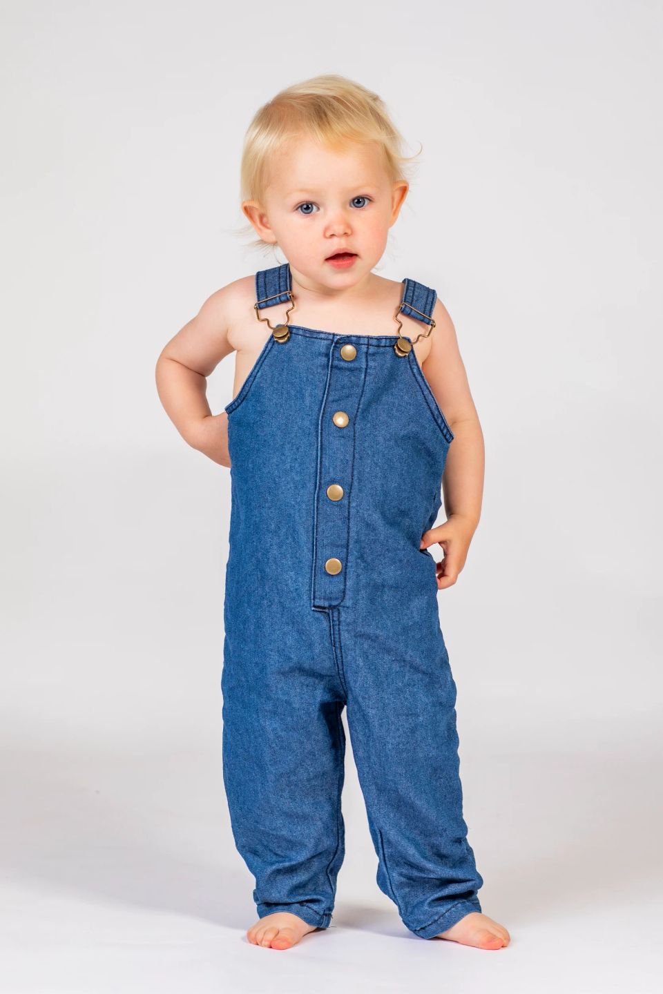 Børn portrætfoto af lille barn med blondt hår og blå øjne i denim-seler, der kigger nysgerrigt mod beskueren.