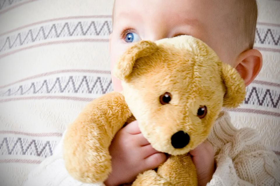 Nærbillede af baby, der holder en lille bamse op til ansigtet med store, blå øjne kiggende frem.