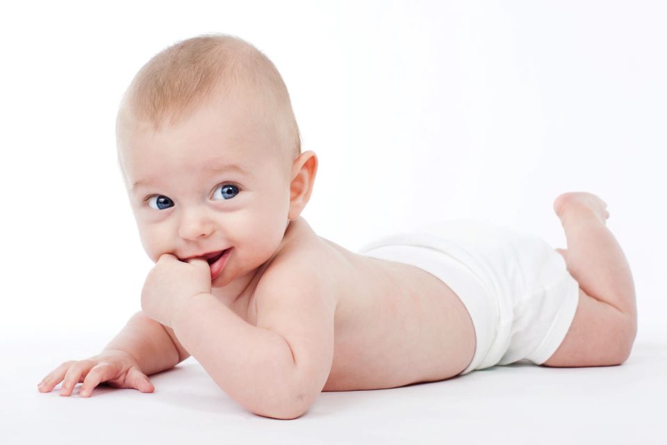 Autentisk baby foto af et smilende barn på maven i hvide blebukser, med blå øjne, kiggende direkte ind i kameraet.