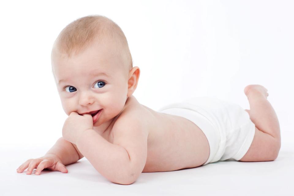 Autentisk baby foto af et smilende barn på maven i hvide blebukser, med blå øjne, kiggende direkte ind i kameraet.
