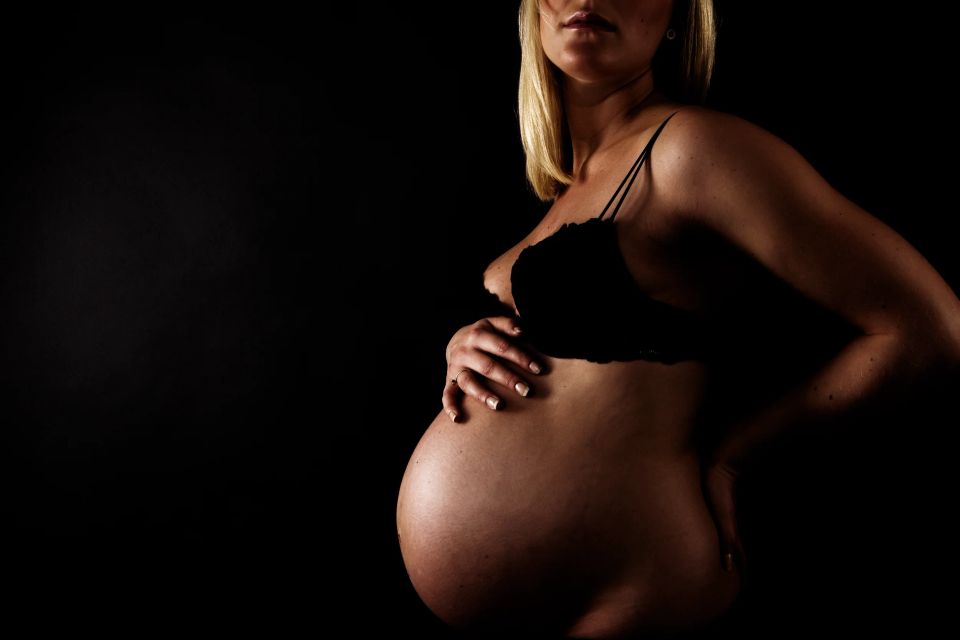 Gravid kvinde i sort bh med hånden på sin gravide mave i dramatisk lys mod mørk baggrund. Graviditetsfotografering.