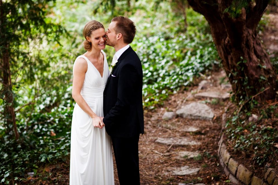 Bryllupsfoto af brudepar i grønne omgivelser, hvor brudgommen kysser bruden på kinden. De holder hånd og smiler.