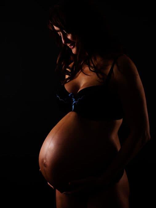 Gravid kvinde i profil, belyst af sidelys der fremhæver hendes mave mod den mørke baggrund. Stemningsfuldt gravidfoto.