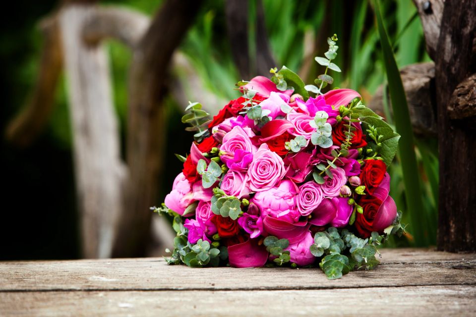 Farverig brudebuket med røde og pink roser, pæoner og eukalyptus på et rustikt træbord. Bryllupsfoto i blødt lys.