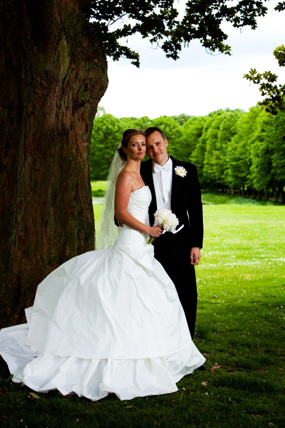 Bryllupsfoto af brudepar under et stort træ i blødt lys, omgivet af græs og grønne træer i baggrunden.