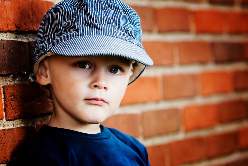 Nærbillede af dreng med stribet kasket, der kigger direkte ind i kameraet, op ad en varm murstensvæg. Enkelt og stemningsf...