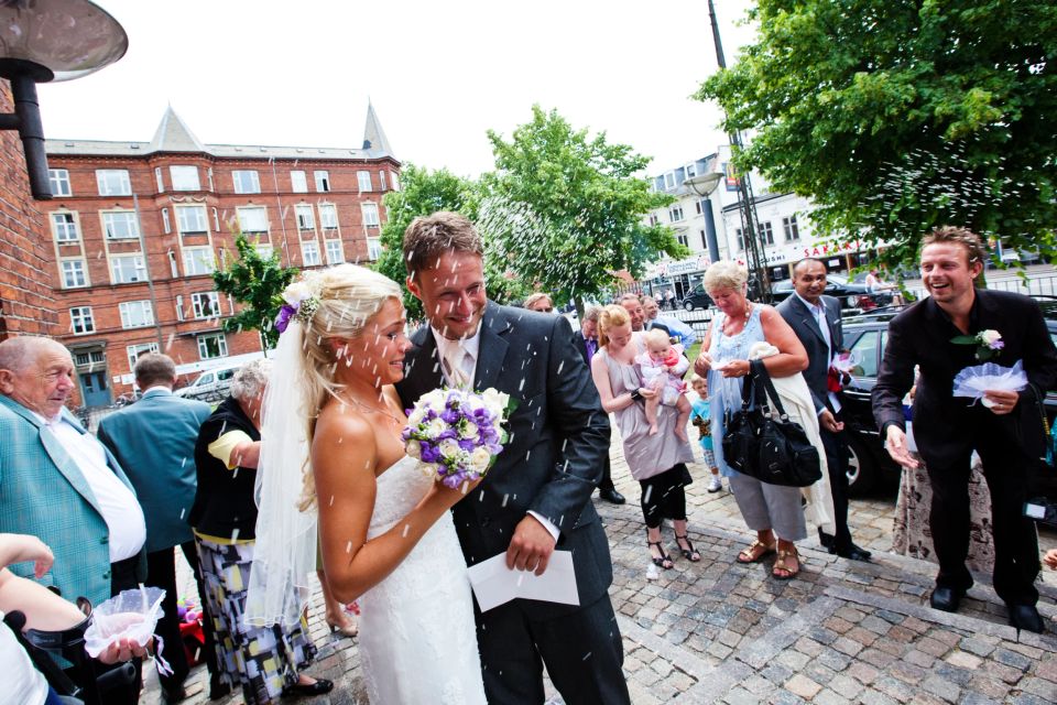 Bryllupsfoto i sorthvid af brudepar dækket af ris uden for en kirke, omgivet af gæster i Virum. Brudeparret smiler.