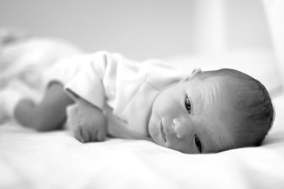 Sort/hvidt foto af en nyfødt baby, der ligger på maven på et hvidt underlag og kigger opmærksomt mod højre. Et fint “andet...