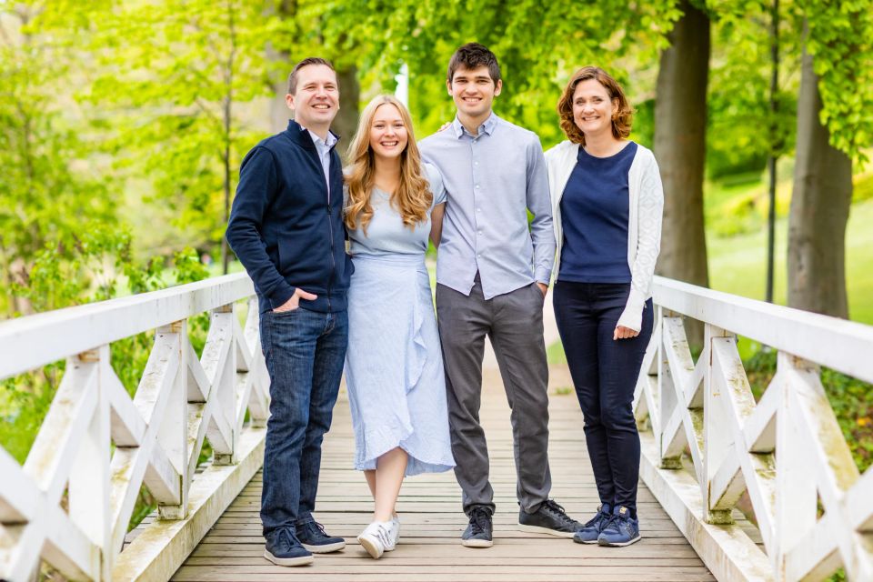 Familiefoto af fire smilende personer på en hvid træbro i solrigt grønt landskab. Et naturligt familieportræt.