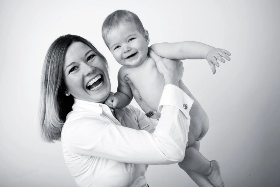 Glad mor løfter sin leende baby op i luften, tæt på i sort/hvid med skarp belysning. Et dejligt babybillede.