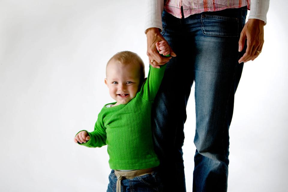Glad baby i grøn trøje, der smiler og holder fast i mors hånd. Et livligt børnefoto med høj kontrast.