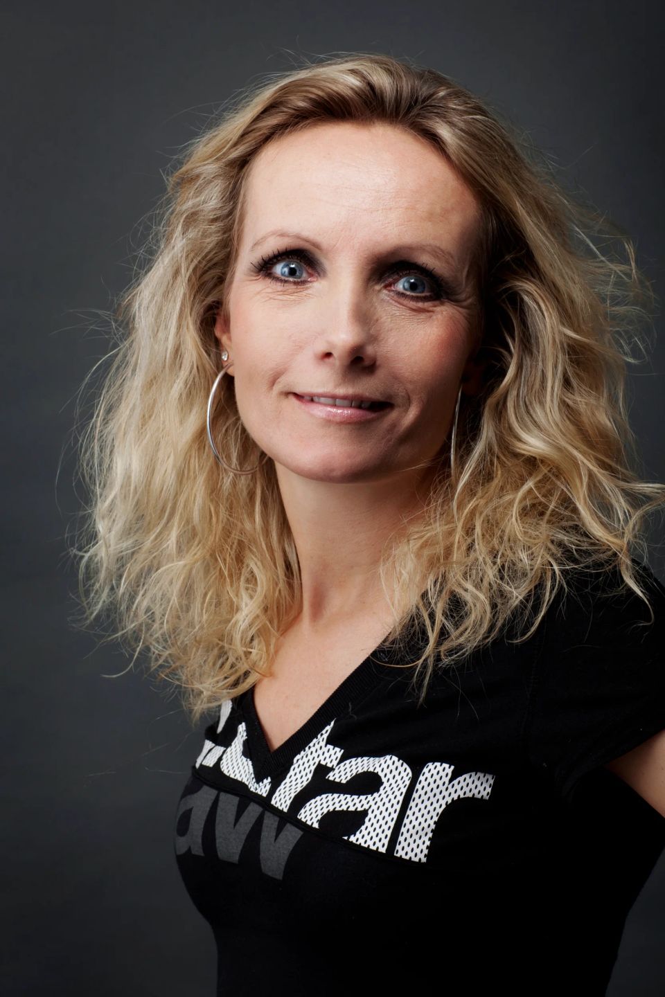Portrætfoto af smilende blond kvinde med krøllet hår og øreringe, iført sort t-shirt mod neutral baggrund i Virum.