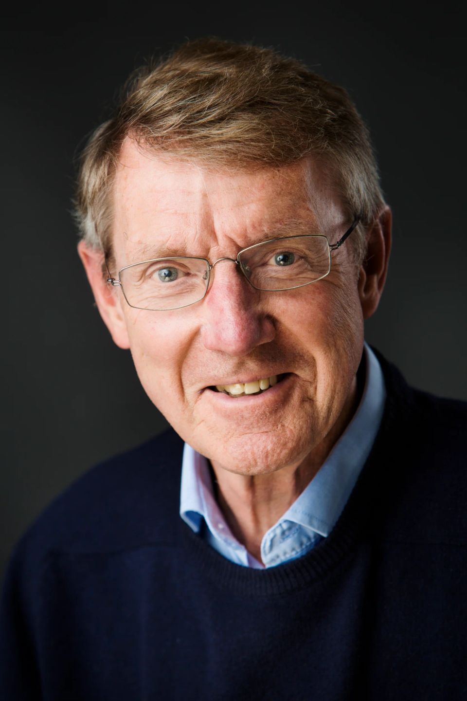 Portrætfoto af ældre mand med briller og et varmt, autentisk smil mod en mørkere, blød baggrund.