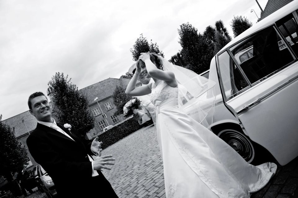 Bryllupsfoto i s/h af brudeparret der netop er ankommet i bil og griner, med bruden der holder sløret på plads.