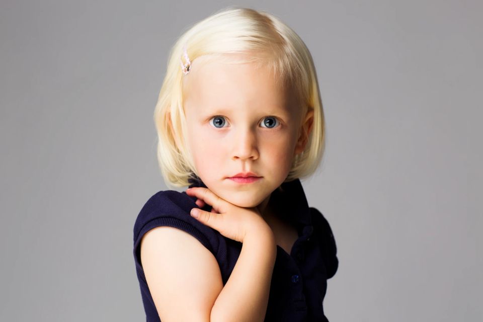 Portrætfoto af lille pige med lyst, kort hår og blå øjne, der hviler hånden ved halsen med grå baggrund.