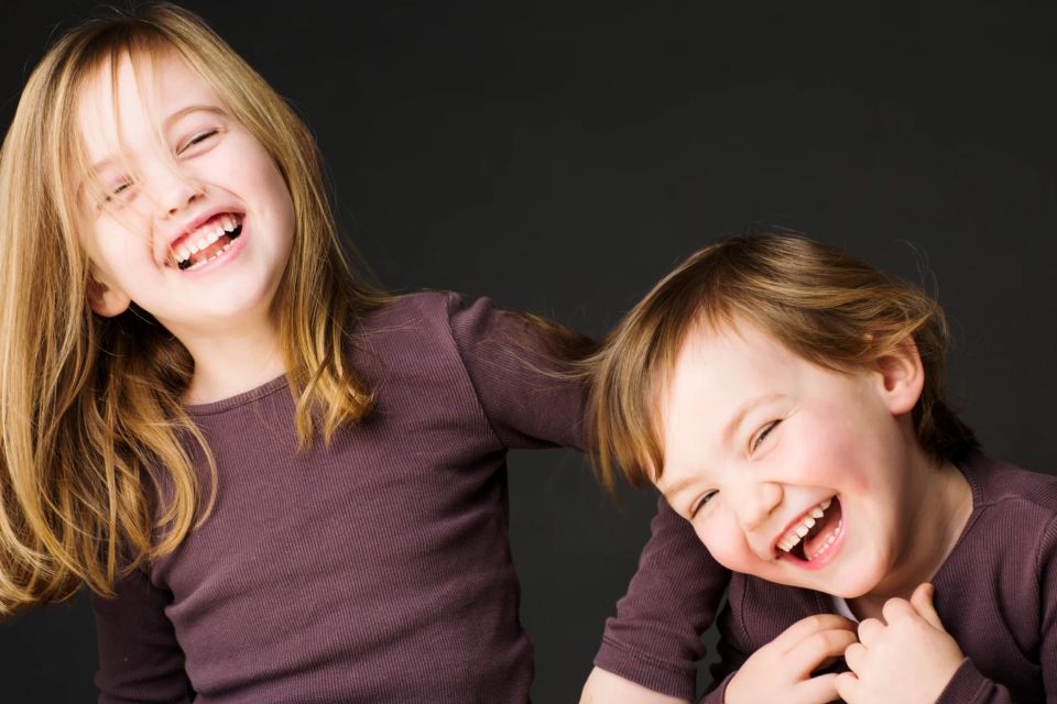 Glade søskende - to børn med lyst hår, der griner hjerteligt og smittende mod mørk baggrund. Et livligt portrætfoto.