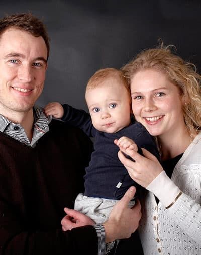 Hjertevarmt familieportræt med smilende forældre og en baby, der kigger nysgerrigt i kameraet. Fotografering af glæde.