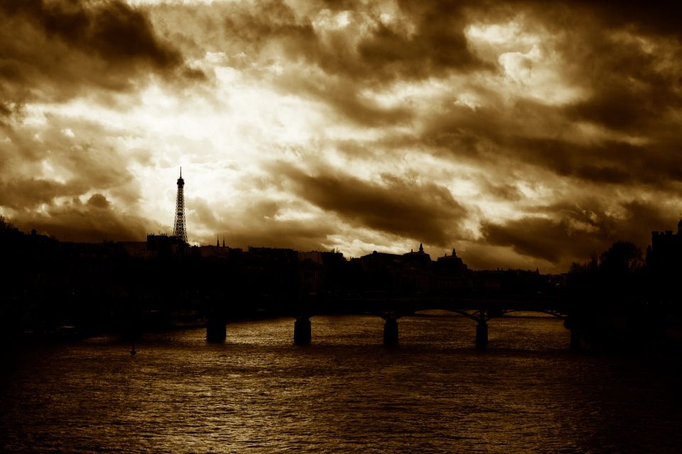 Stemningsfuldt foto af Eiffeltårnet og Seinen i Paris under en dramatisk mørk himmel med gulligt lysbrydning. En bro ses o...