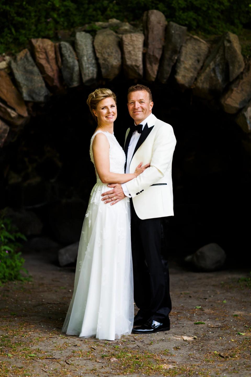 Bryllupsfoto af smilende par i hvid brudekjole og hvid smoking stående foran en mørk grotteåbning i dagslys.