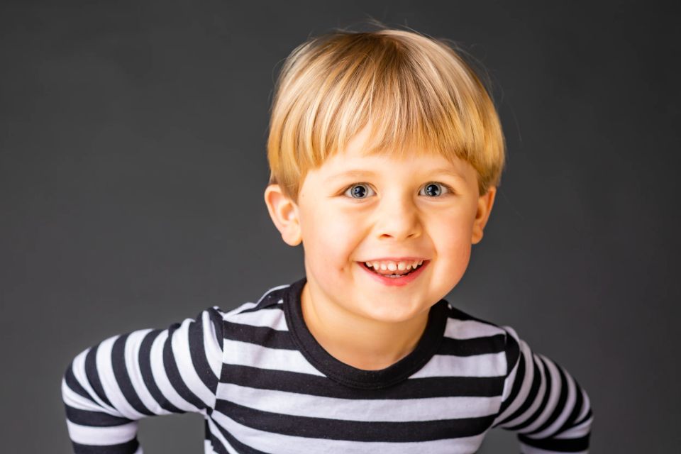 Børnefoto af en glad lille dreng med blond hår og store, lyse øjne, der smiler bredt mod et mørkt bagtæppe.