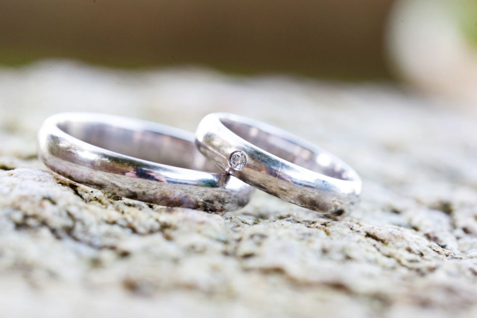 Nærbillede af to skinnende sølv vielsesringe, den ene med en lille diamant, liggende på en lys sten. Bryllupsfotografi.