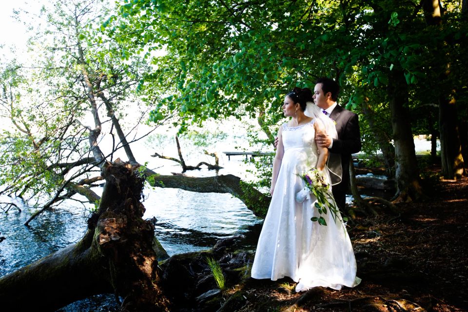 Bryllupsfotografering: Brudepar står ved søbred. Grønne træer, vand og flydende træstamme bag parret i varmt lys.