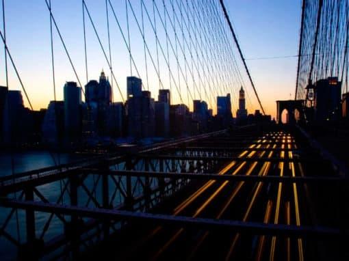 Bro og New York skyline i skumring. Aftenhimlen spejles i sorte bygningsfacader og vand. Lange lysstriber fra biler.