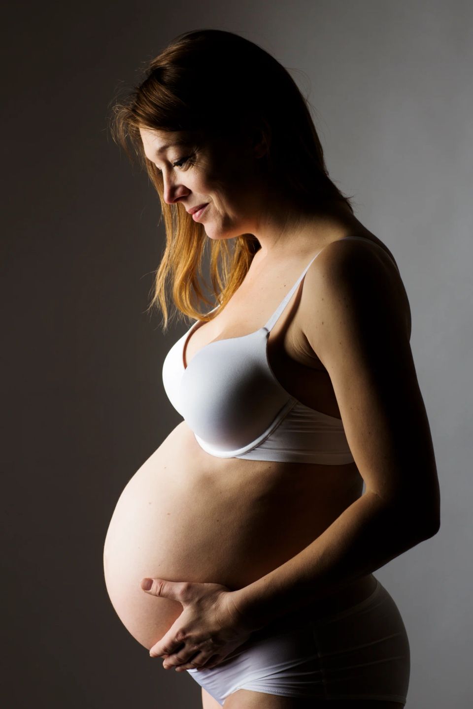 Gravid kvinde fotograferet i profil, iført hvidt undertøj, holder om sin mave i blødt sidelys. Graviditetsfotografering.