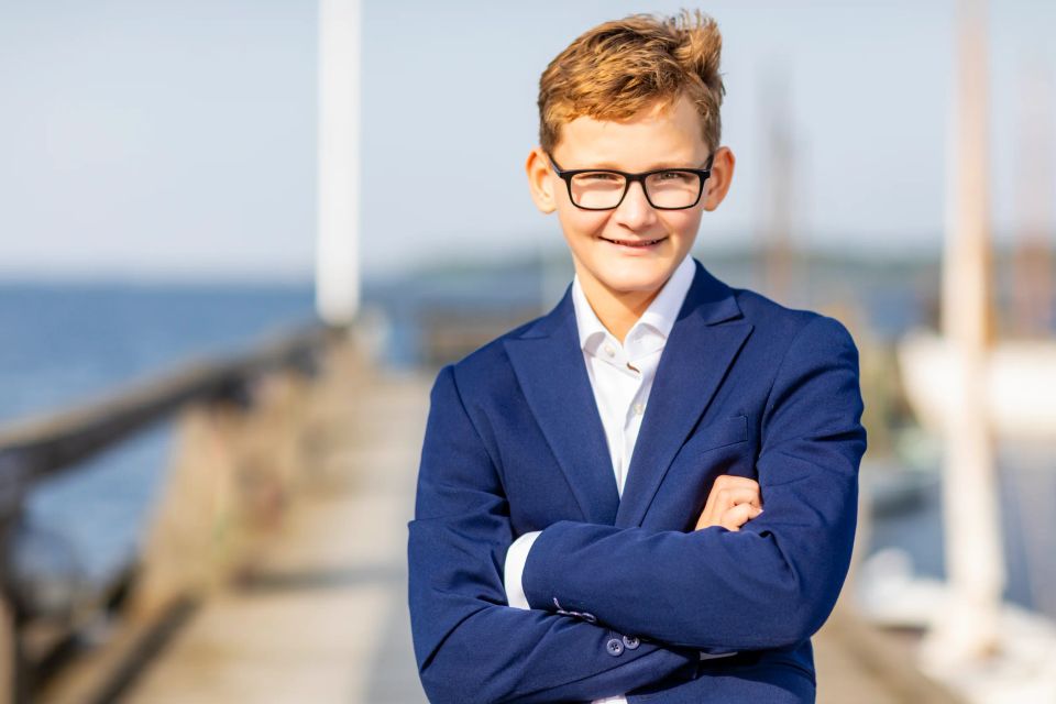 Portrætfoto af glad konfirmand i mørkeblåt jakkesæt med armene over kors på en solrig havn i Virum.