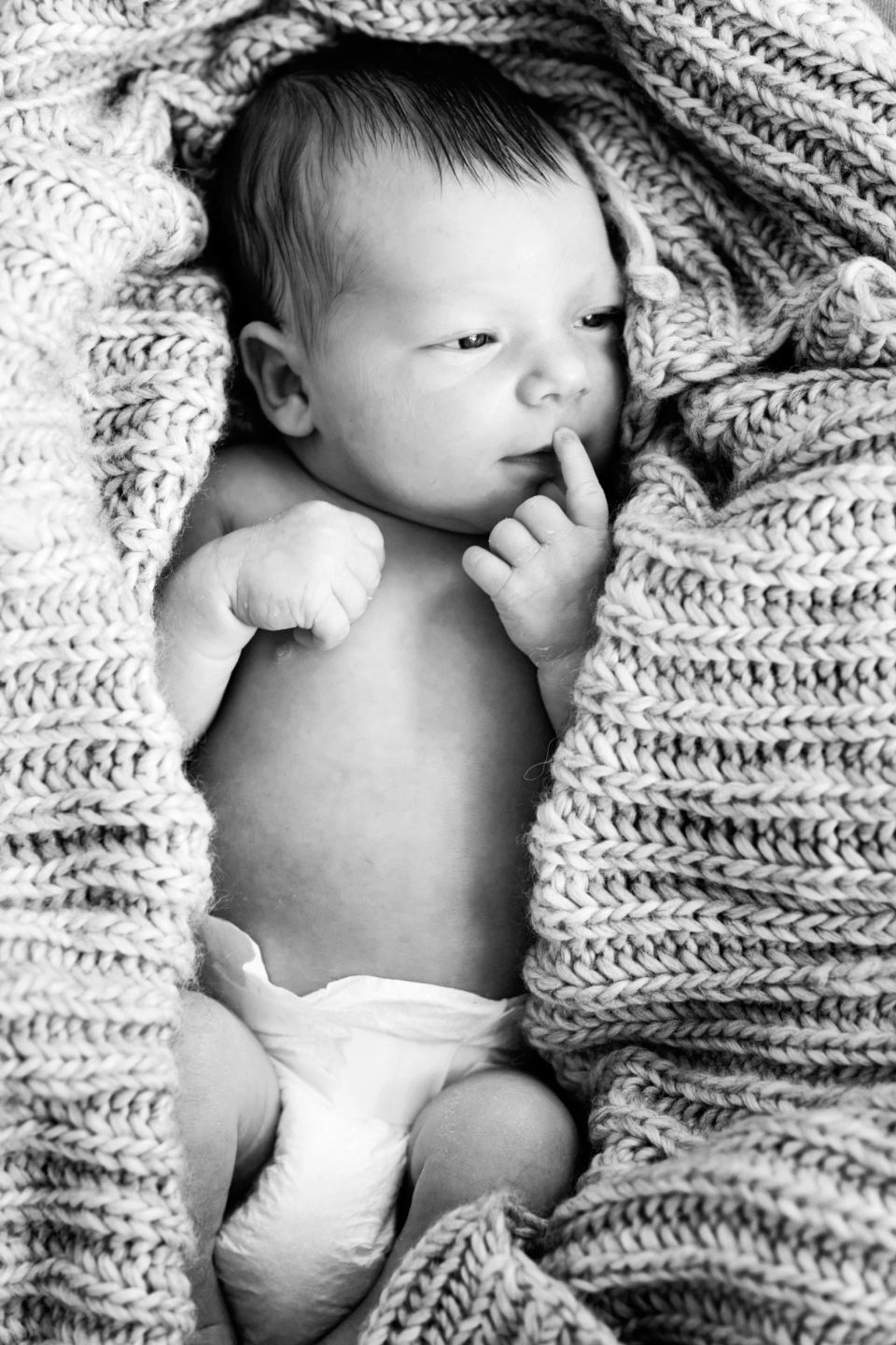 Sort/hvid babyfoto: Nyfødt puttet i blødt tæppe, finger ved mund, let åbne øjne, fredfyldt stemning.